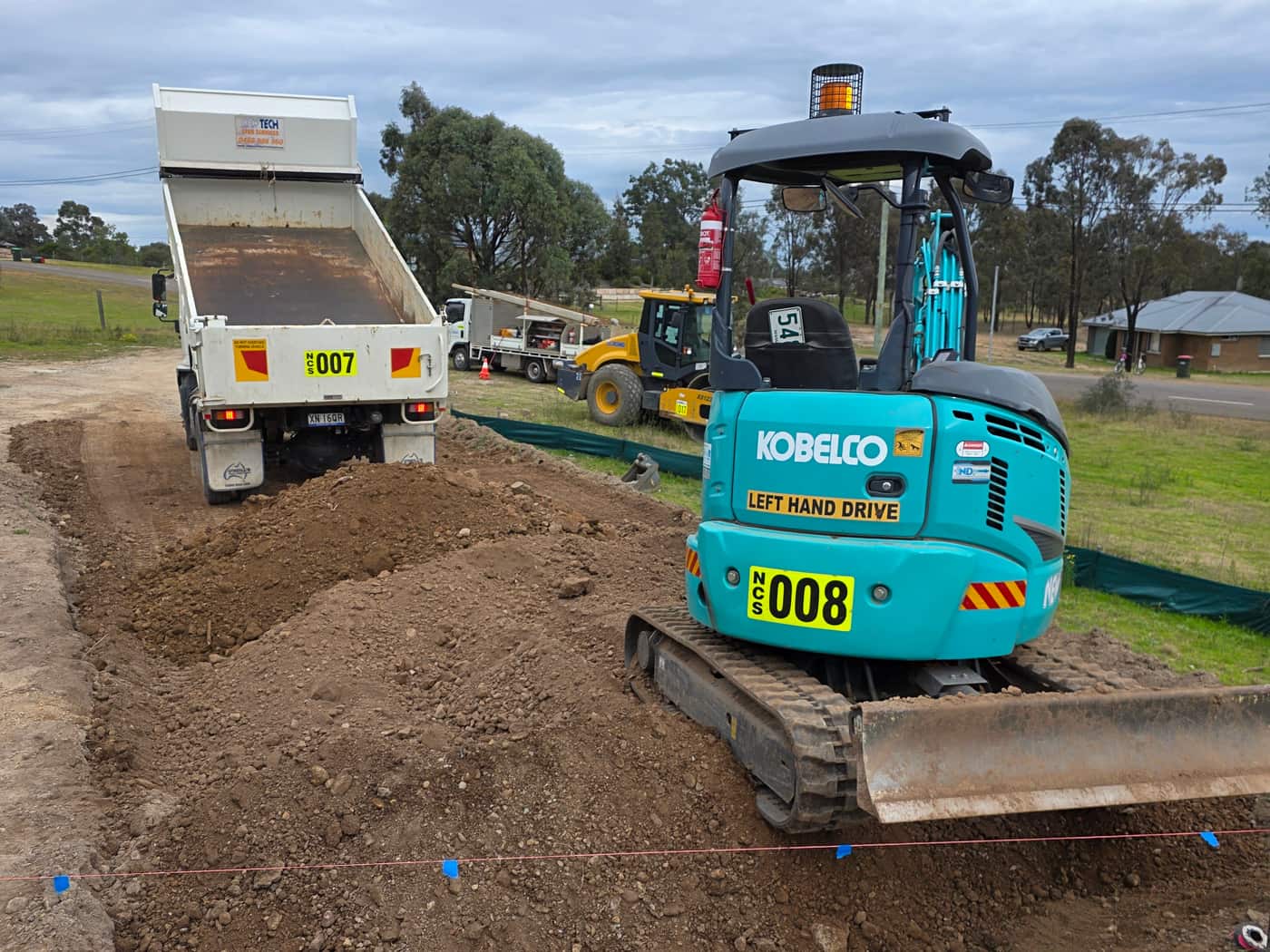 A Newtech mini excavator cutting a pad with the tipper backed in behind it and string lines pegged out in front