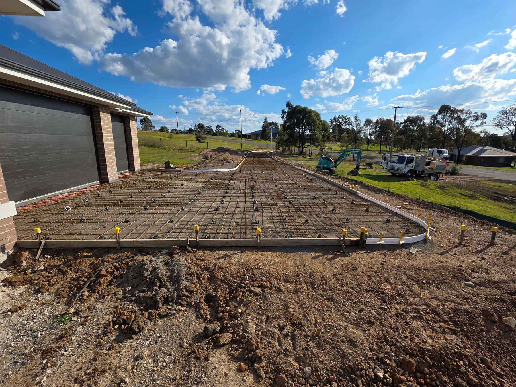 A long slab boxed up and meshed beside a new house, with the excavator and truck on the block behind